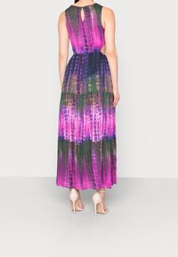 Robe longue multicolore avec un motif tie-dye vibrant en violet, vert et bleu. Le dos comporte un détail en forme de trou de serrure. Portée avec des talons beiges.