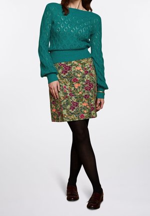 Femme portant un pull texturé bleu sarcelle, une jupe fleurie longueur genou, des collants noirs et des mocassins marron, debout les jambes croisées sur fond blanc.