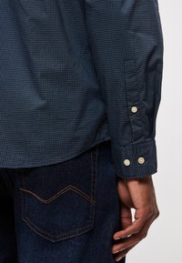 Μπλε και τιρκουάζ καρό πουκάμισο με κουμπιά στους μανικιών, συνδυασμένο με σκούρα τζιν με αντίθετο σχέδιο ραφής στην τσέπη.