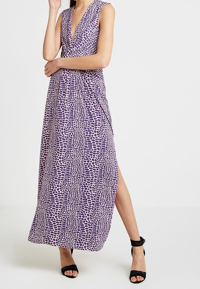 Femme portant une robe maxi violette sans manches à motif, avec un décolleté en V profond et des sandales noires à talons hauts, debout avec une jambe visible.