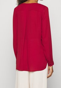 Blusa rossa a maniche lunghe realizzata in tessuto leggero. Presenta un colletto arrotondato, una piega sul retro e polsini con bottoni. Texture liscia.