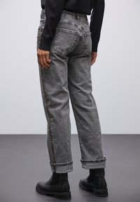 Jean en denim gris avec une coupe décontractée, ourlets retroussés et une couture décorative en or sur le côté. Porté avec des bottines chelsea noires.
