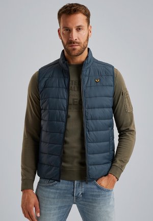 Gilet imbottito trapuntato blu navy con collo alto, chiusura con zip e piccolo logo sul lato sinistro del petto, indossato sopra una camicia a maniche lunghe color oliva e jeans blu.
