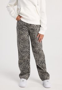 Person wearing white hoodie, leopard print wide-leg pants, and white sneakers, standing with one hand in pocket against plain background.