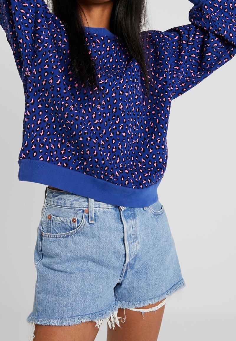 Person wearing a blue sweatshirt with pink and black leopard print and light blue frayed denim shorts, arms raised above head.