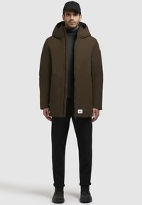 Mørkebrun hætteparka med frontlynlås, to sidelommer og et mærke ved kanten. Båret over en sort rullekrave og bukser.