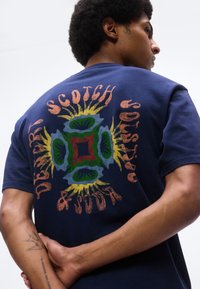 Mand med korslagte arme iført mørkeblå t-shirt med farverigt abstrakt blomsterdesign og ordene "Scotch & Soda Denim" på bagsiden.