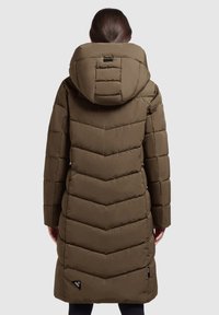 Naine kannab pikka pruuni tikitud puffjakki koos suure kapuutsi ja seismisega neutraalse tausta ees, seljaga pöördununa.