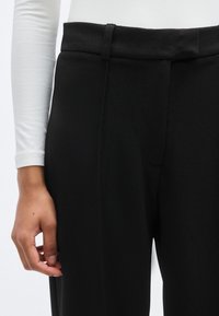 Μαύρο παντελόνι με λείο ύφασμα και εφαρμοστή γραμμή. Διαθέτει επίπεδη μέση και λεπτομέρειες στις πλευρικές ραφές. Χέρι τοποθετημένο πάνω στο ένδυμα.