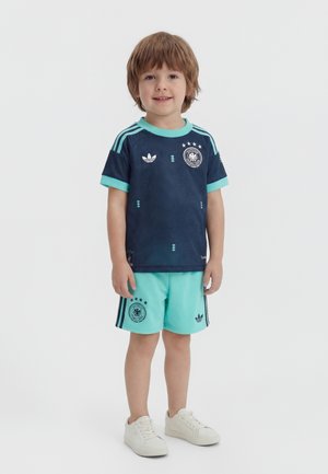 Malé dieťa stojace, oblečené v tmavomodrom a tyrkysovom športovom drese a zodpovedajúcich šortkách s bielymi teniskami na jednoliatom pozadí.