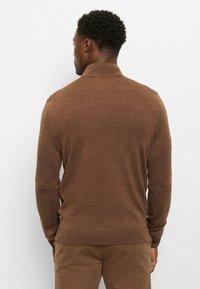 Brauner langärmeliger Pullover mit strukturierter Strickoptik, ausgestattet mit einem Kragen und einem geraden Saum, getragen zu khakifarbener Hose in neutraler Umgebung.