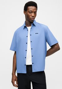 Homme portant une chemise bleu clair à manches courtes boutonnée par-dessus un t-shirt blanc, associée à un pantalon noir, debout devant un fond uni.