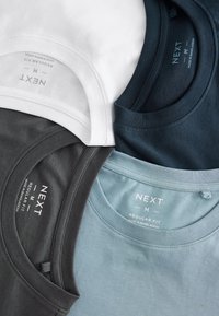 Quattro t-shirt girocollo piegati in bianco, nero, blu navy e azzurro chiaro, ognuno etichettato come medium regular fit del marchio NEXT.