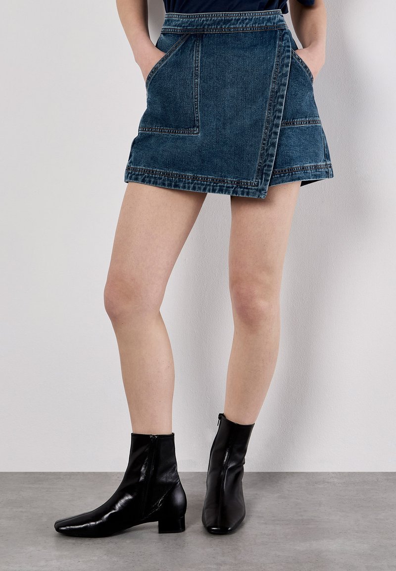Jupe portefeuille en denim de lavage foncé avec deux poches avant, associée à des bottes en cuir noir à bout carré et à petit talon carré.