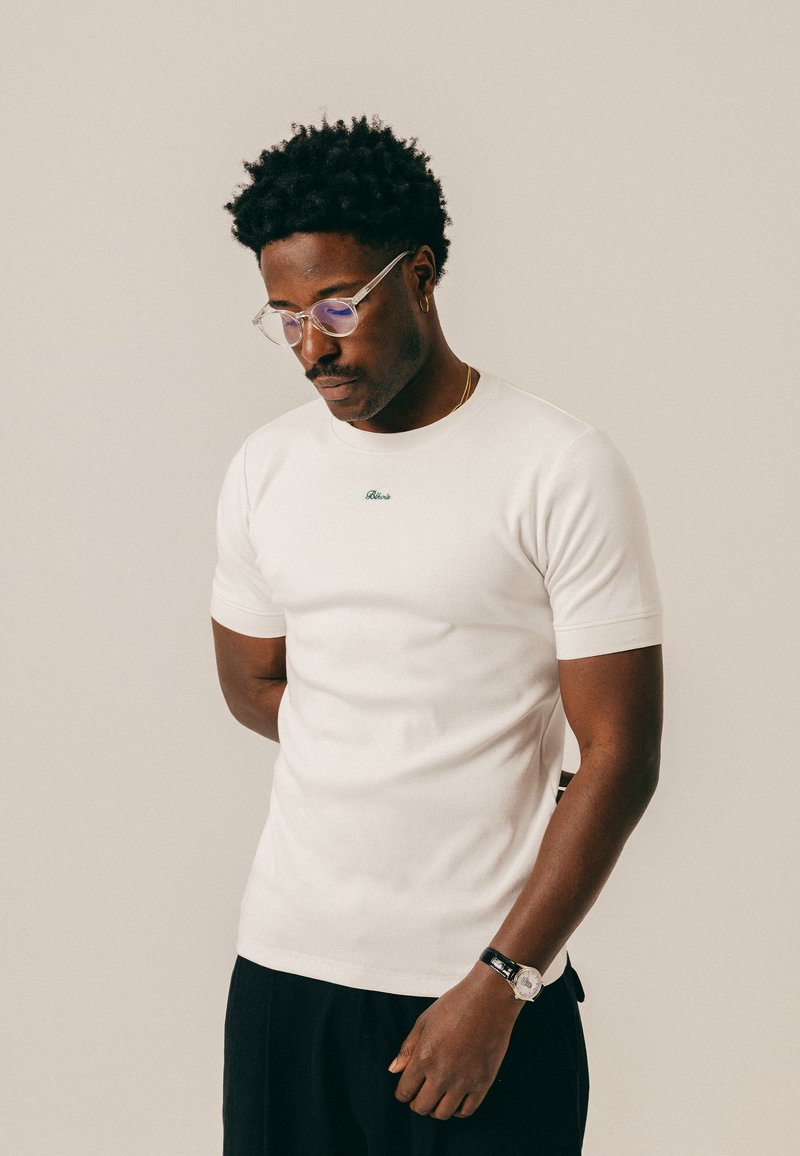 Άνδρας με κοντά σγουρά μαλλιά και μουστάκι φορά διαφανή γυαλιά, λευκό μπλουζάκι, μαύρο παντελόνι, χρυσό κολιέ, σκουλαρίκι κρίκο και ρολόι χειρός, κοιτάζει προς τα κάτω.