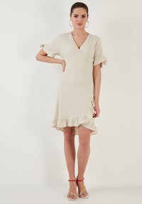 Wrap dress in light beige with a textured finish, short sleeves, ruffled hem, and side tie detail, paired with multicolored sandals.