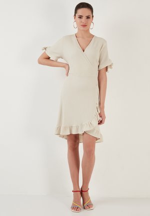 Wrap dress in light beige with a textured finish, short sleeves, ruffled hem, and side tie detail, paired with multicolored sandals.