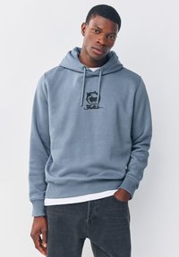 Szara bluza z kapturem z kieszenią typu kangur, z czarnym logo i sznurkami. W zestawie z białą koszulką i ciemnymi dżinsami. Miękka w dotyku.