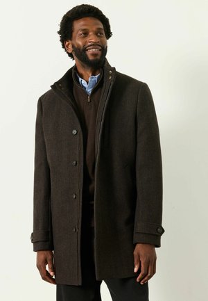 Mann mit Bart trägt einen dunkelbraunen, geknöpften Mantel über einem braunen Zip-up-Pullover und einem blauen Hemd mit Kragen, lächelt und schaut zur Seite.