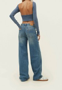 Kvinna i en långärmad crop top med öppen rygg, bredbeniga blå jeans och vita sneakers, stående vinklad bort mot en ljus bakgrund.