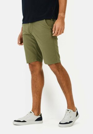 Olive green shorts made of cotton blend, feature side pockets, flat front, and a relaxed fit. Worn with white sneakers.