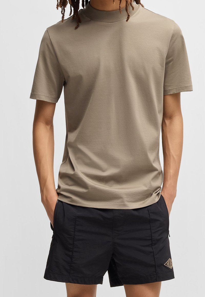 Personne portant un t-shirt beige uni à manches courtes et un short noir, les mains dans les poches, debout sur un fond uni.