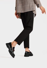 Mocassini in pelle verniciata nera con suola spessa, dotati di hardware decorativo. Indossati con pantaloni neri su misura e un blazer a quadri.