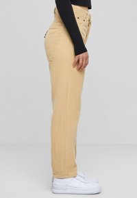 Pantaloni in velluto a coste color beige chiaro con vestibilità dritta, caratterizzati da un alto punto vita, tasche laterali e un accento colorato sulla parte posteriore. Sneakers bianche.
