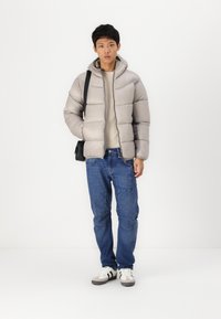 Lichte grijze gewatteerde jas met horizontale stiksels, gecombineerd met een beige shirt en blauwe spijkerbroeken. Voorzien van een capuchon en zakken.