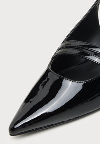 Chaussure en cuir verni noir brillant à bout pointu avec deux brides décoratives sur le dessus, présentée sur un fond clair uni.