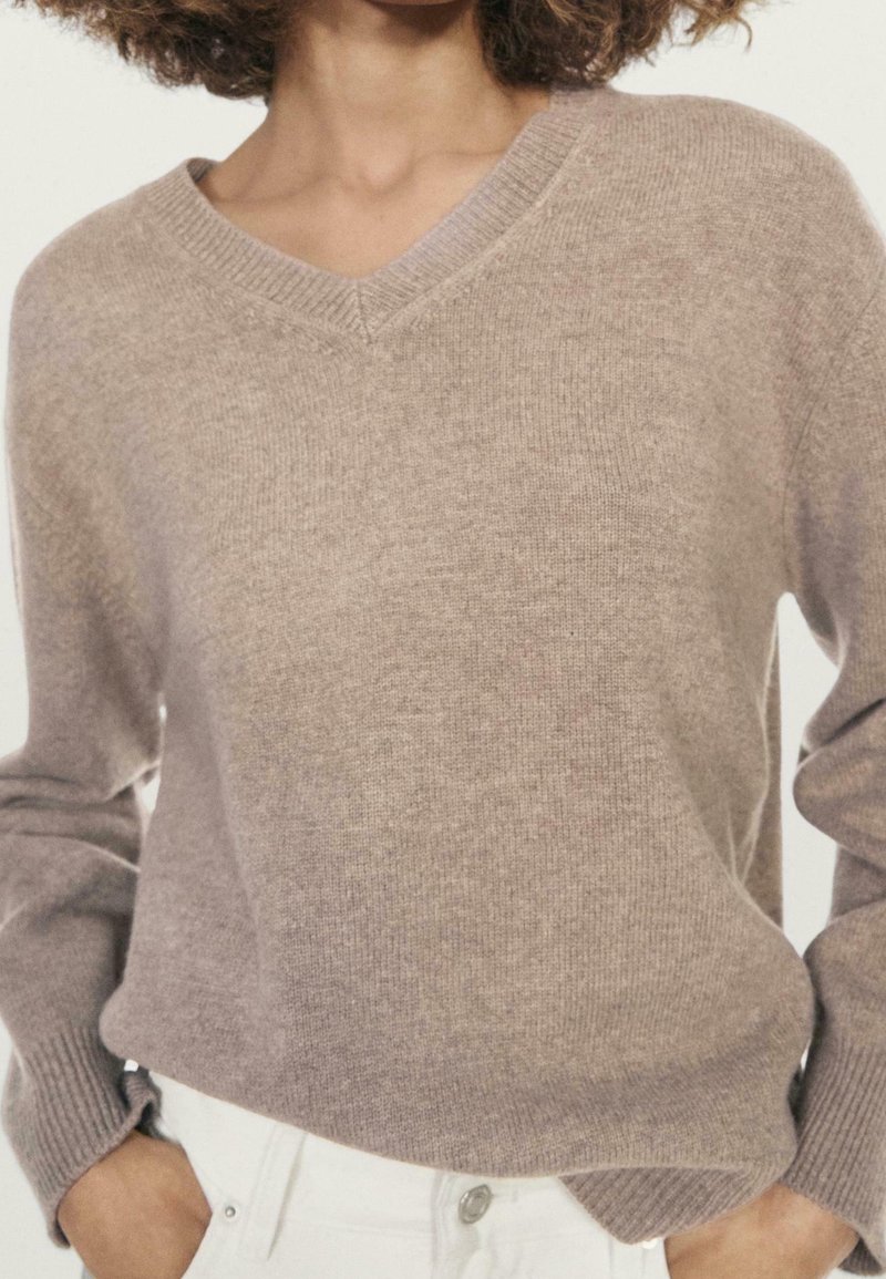 Femme portant un pull en maille taupe col en V avec des poignets côtelés, rentré dans un pantalon clair, mains partiellement dans les poches.