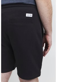 Sorte bomullsshorts med ribbestrikket liv, som har en enkelt baklomme og en liten hvit merkeetikett på høyre side.