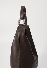 Hnědá kožená kabelka s texturovaným povrchem, jediným popruhem upevněným stříbrným kovovým doplňkem, zúženým tvarem a bez viditelného zapínání.
