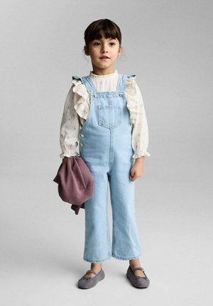 Lichtblauwe denim overall met brede pijpen, met daaronder een witte blouse met ruches en lange mouwen. Grijze schoenen en een zacht paars sweater in de hand.