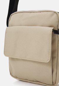 Borsa a tracolla in tessuto beige con tasca frontale a patta, forma rettangolare, dettagli cuciti e tracolla nera regolabile.