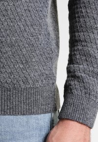 Grauer Strickpullover mit einem strukturierten Rautenmuster, gerippten Bündchen und Saum. Hergestellt aus weichem, warmem Stoff; das Detail zeigt einen Teil des Arms und der Hand.