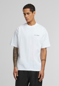 White cotton t-shirt featuring short sleeves, a crew neck, and a small blue embroidered logo on the left chest area. Simple, relaxed fit.
