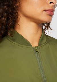 Zelená bomber bunda s hladkou texturou, žebrovaným límcem a zipovým předním zapínáním. Disponuje jednoduchým designem bez dalších vzorů či akcentů.