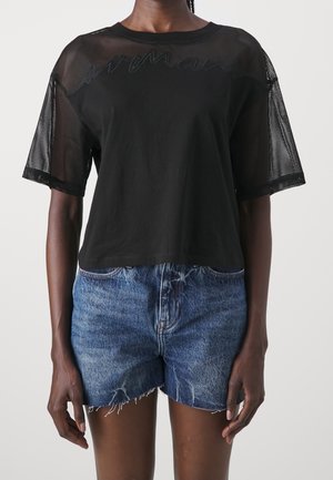 Nainen, jolla on musta lyhythihainen verkkokankainen ja sileä kangastoppi, yhdistettynä korkeavyötäröisiin, sorvatun reunan sinisiin farkkushortseihin.