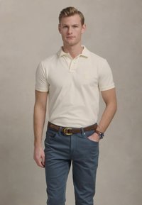 Kremo spalvos polo marškiniai su apykakle ir trumpomis rankovėmis, derinami su mėlynomis kelnėmis. Marškiniai turi nedidelį logotipą krūtinės srityje ir rudą diržą.