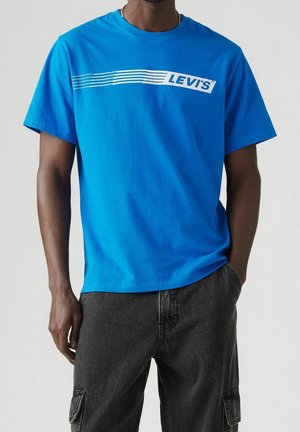 Mand iført en klarblå Levi's T-shirt med hvidt logo og horisontale striber, kombineret med sorte cargobukser.