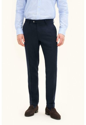Man draagt marineblauwe op maat gemaakte broek, lichtblauw gestreept overhemd en donkerbruine loafers, staat rechtop met ontspannen handen.