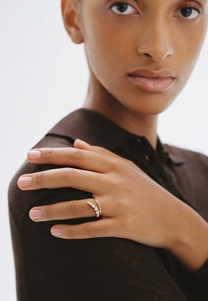 Mujer con una camisa oscura muestra su mano con un delicado anillo de oro que tiene una fila curva de pequeños diamantes sobre su hombro.