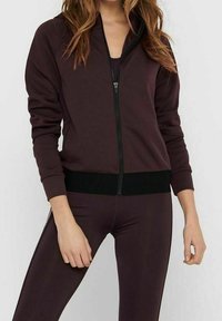 Burgundská zipová bunda s černým žebrovaným lemem. Vyrobena z měkkého materiálu, má vysoký límec a přiléhavý střih. Nosená přes odpovídající legíny.