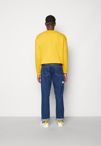 Gul sweatshirt med sänkta axlar, blå raka jeans och vita sneakers med gula accenter, sedd bakifrån.