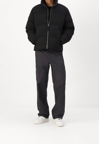 Osoba w czarnej pikowanej kurtce, ciemnoszarych spodniach typu cargo i białych trampkach stoi z rękami w kieszeniach na białym tle.