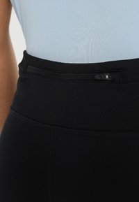 Gros plan sur une personne portant un legging noir taille haute avec une petite poche zippée horizontale sur la taille et un haut bleu clair.