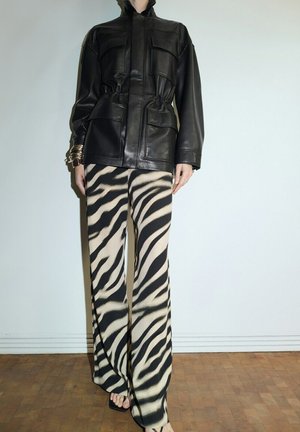 Persona che indossa una giacca di pelle nera con quattro tasche frontali e pantaloni a gamba larga a righe zebra su un pavimento di legno.