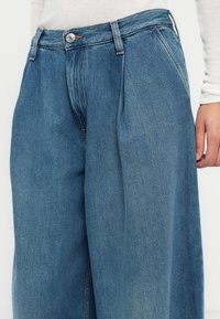 Pantalon large en denim bleu à taille haute, avec plis devant, bouton argenté et tissu texturé.