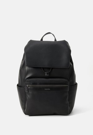 Sac à dos en cuir noir avec un rabat en haut, une poche frontale zippée et des poches latérales. Il est doté d'une poignée en boucle et d'une texture extérieure lisse.
